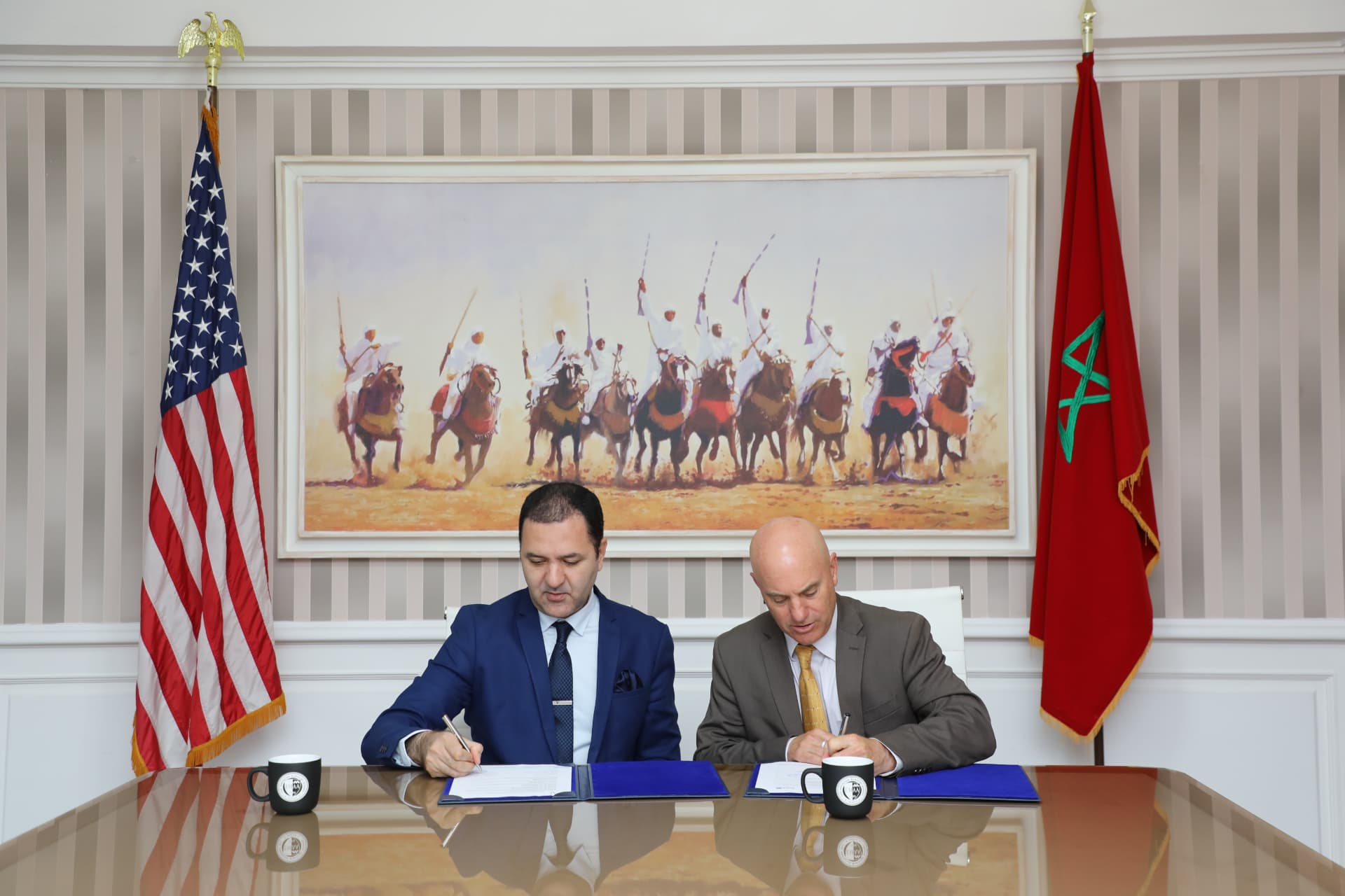 Two officials in suits signing agreement documents at a table flanked by the United States and Moroccan flags