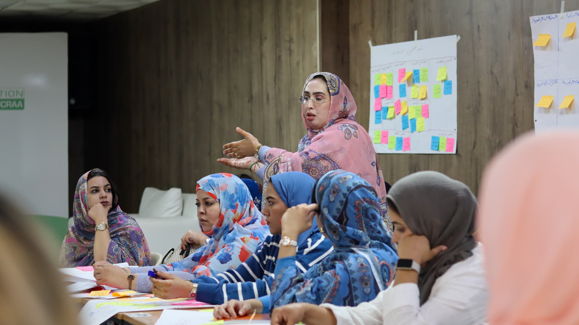 A facilitator addressing seated participants in front of a wall covered with sticky notes