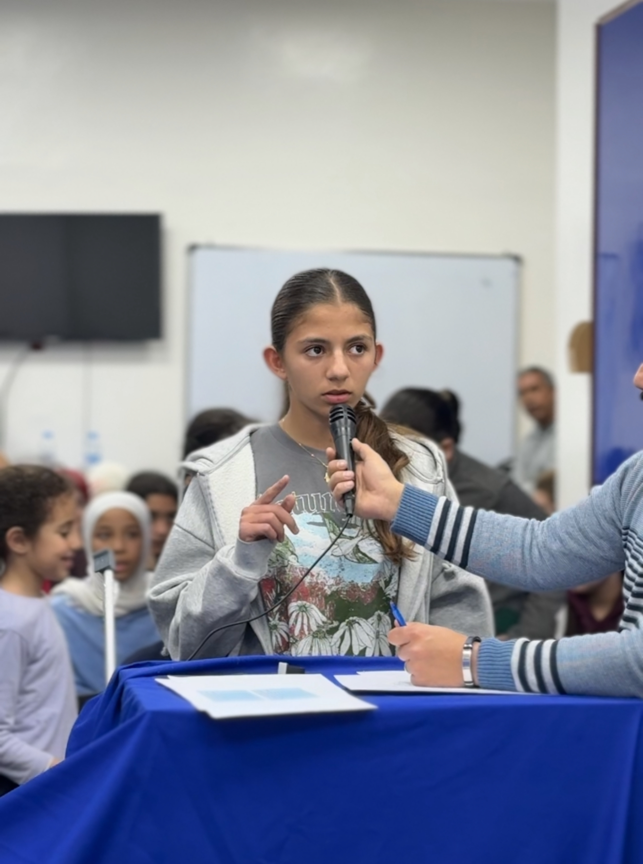 A young student speaking into a microphone held by a host at a blue-draped podium