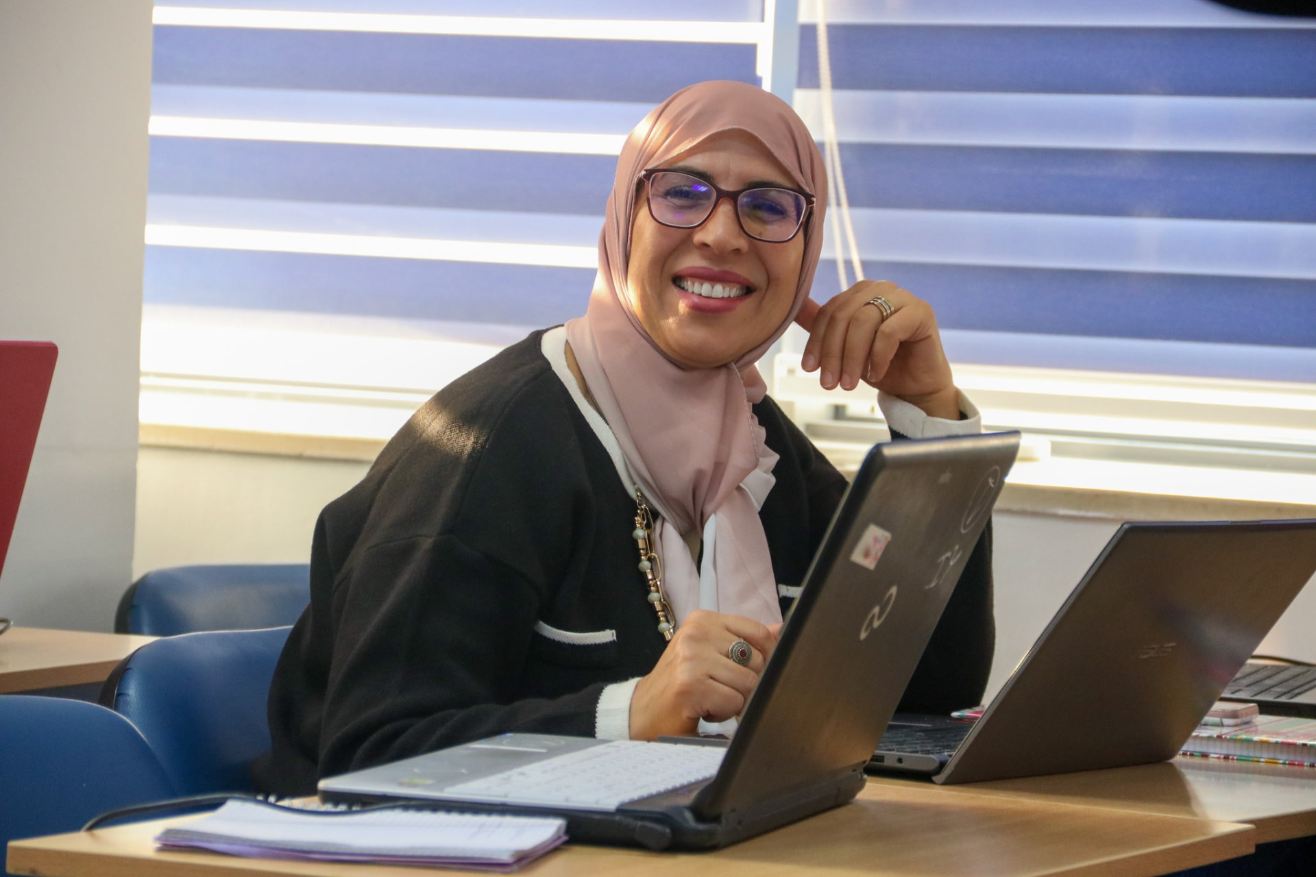 A woman smiling at the camera while working at a desk with two open laptops